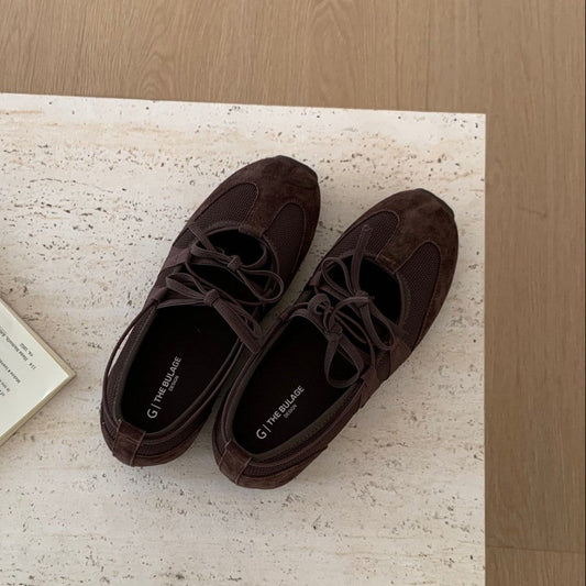 A pair of dark brown suede lace-up flats with a square toe, styled on a textured beige stone surface next to an open book and a small green sprig. The brand name 'the square.