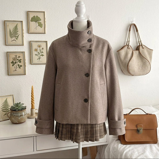 A taupe-brown high-neck wool coat displayed on a mannequin torso, styled with a plaid skirt. The setting is a cozy bedroom with a white dresser, framed botanical prints, a hanging beige tote bag, and a brown leather handbag on a bed.
