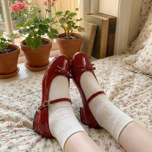Red shoes with bows on a floral bedspread, surrounded by potted plants and books.