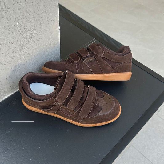 A pair of dark brown suede low-top sneakers with wide hook-and-loop straps and gum soles, displayed on a black platform against a gray tiled floor.