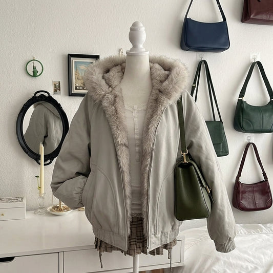 A light gray hooded jacket with a fluffy faux-fur lined hood is displayed on a mannequin torso in a bedroom setting. The jacket is styled over a white top and plaid skirt, with several handbags hanging on the wall behind it.