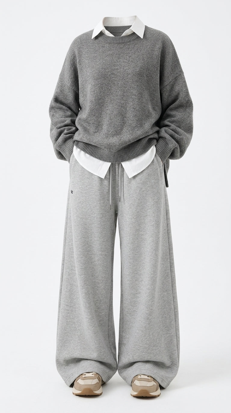 A person wearing a gray knit sweater over a white collared shirt, paired with wide-leg gray sweatpants and beige sneakers, photographed against a plain light gray studio background.