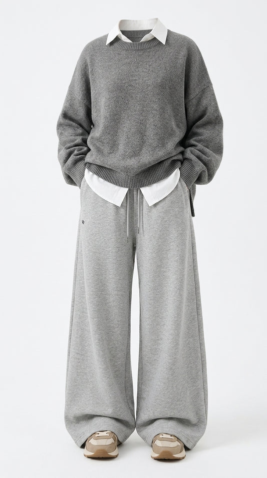 A person wearing a gray knit sweater over a white collared shirt, paired with wide-leg gray sweatpants and beige sneakers, photographed against a plain light gray studio background.