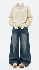 A child wearing a cream knit V-neck sweater over a white collared shirt, paired with wide-leg blue jeans and beige sneakers, shown against a plain light gray studio background.