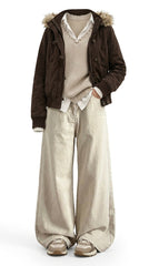 A full-body studio shot of a person wearing a dark brown corduroy hooded jacket with faux-fur trim, a beige knit sweater vest, a white collared shirt, and wide-leg beige trousers, paired with beige and brown sneakers, against a plain white background.