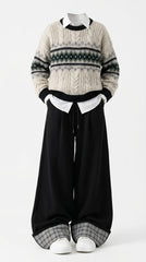 A person wearing a cream cable-knit sweater with black and gray Fair Isle style bands, a white collared shirt underneath, loose black wide-leg pants, and white sneakers, photographed against a plain light gray studio background.