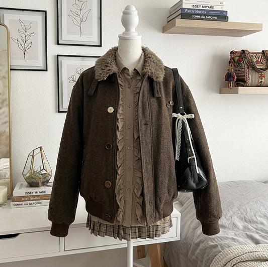 A brown corduroy jacket with a fuzzy collar is displayed on a mannequin in a styled bedroom setting. The jacket is layered over a brown ruffled blouse and a plaid skirt, with a black shoulder bag hanging from the mannequin arm.