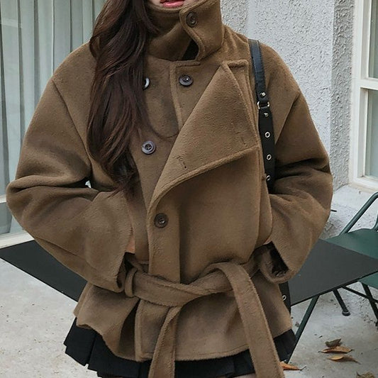 A woman wearing a brown high-neck belted coat with dark tights, standing outdoors in front of a building with windows and a green bench.