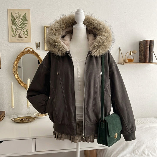 A dark brown faux-leather bomber jacket with a large beige faux-fur trimmed hood, displayed on a white mannequin torso in a bedroom setting. A dark green crossbody bag is draped over the side of the mannequin.