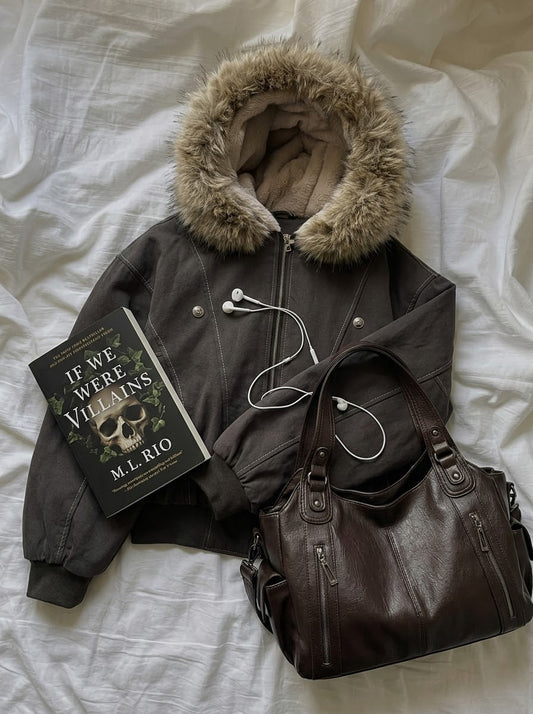 A dark brown faux-fur-trimmed bomber jacket is laid flat on white bedding, styled with a brown leather shoulder bag, a book titled 'If We Were Villains' by M.L.
