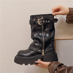 Black leather boot with a gold buckle and zipper held by a hand against a neutral background