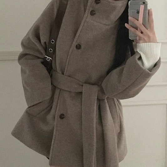 A woman takes a mirror selfie wearing a long taupe-brown belted coat over a white ribbed sweater. The coat features a high mock neck, large front buttons, and oversized sleeves.