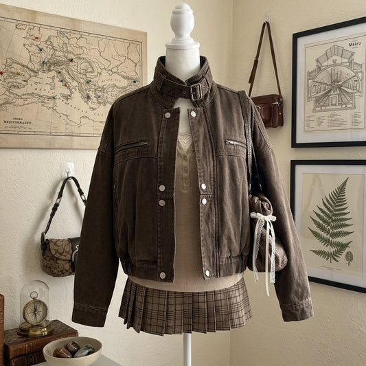 A brown cropped denim jacket displayed on a white mannequin torso, styled with a beige knit top and a brown plaid pleated skirt, set against a vintage-themed room background with framed maps and botanical prints.
