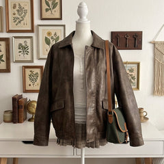 A brown leather jacket displayed on a white mannequin torso, styled with a white top, a plaid skirt, and a green shoulder bag with brown leather straps. The background shows a white wall decorated with framed botanical prints, vintage books, and a macrame wall hanging.