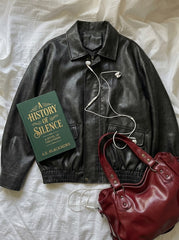 A black leather jacket laid flat on a white fabric background, styled with a green book titled 'A History of Silence' by A.L.