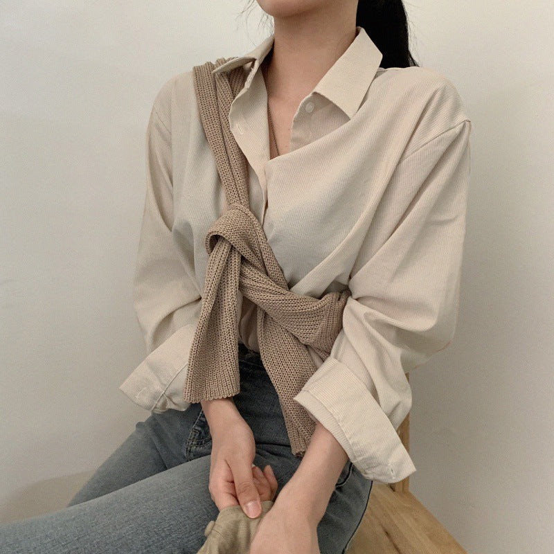 Person wearing a beige shirt with a knitted brown sweater over one shoulder against a plain background