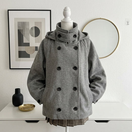 A gray hooded double-breasted coat displayed on a white mannequin torso, set against a white wall with framed art, a round mirror, and a white table with small decor objects.