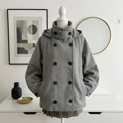 A gray hooded double-breasted coat displayed on a white mannequin torso, set against a white wall with framed art, a round mirror, and a white table with small decor objects.