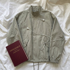 A light olive-gray utility jacket is laid flat on white bedding, styled with a burgundy book titled 'MIDDLEMARCH' and white wired earbuds.