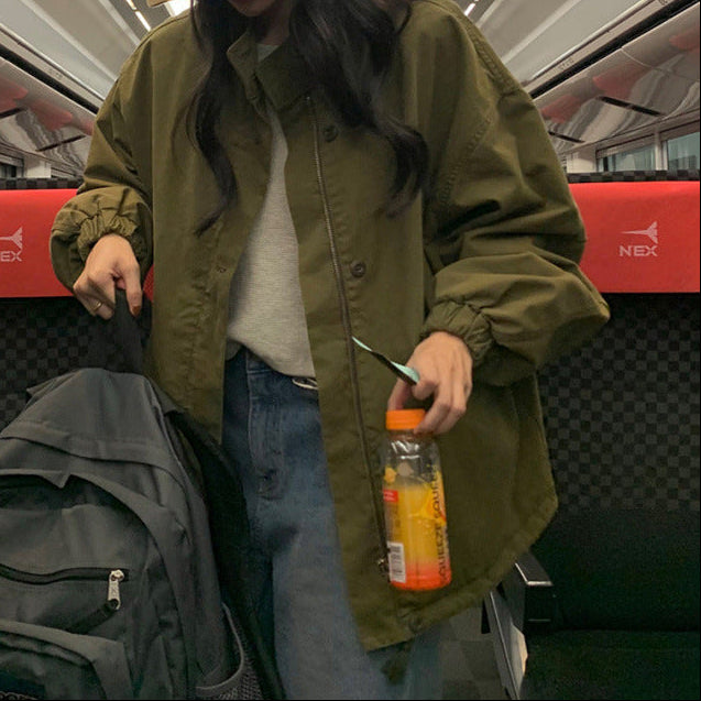 A person standing on a train wearing an olive green utility jacket over a light gray top and blue jeans, with a tan cap. They are holding a clear water bottle and standing next to a gray backpack and a red-and-black seat.
