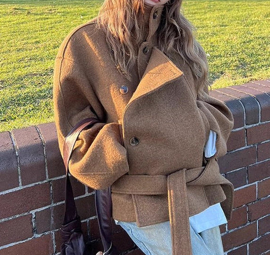A woman wearing a camel brown belted coat, beige pants, and a beige baseball cap leans against a brick wall in an outdoor park setting.