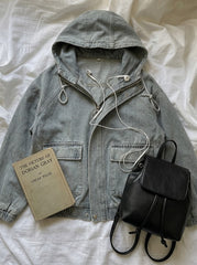 A light gray hooded jacket laid flat on white fabric, styled with a beige book, white earphones, and a black leather backpack.