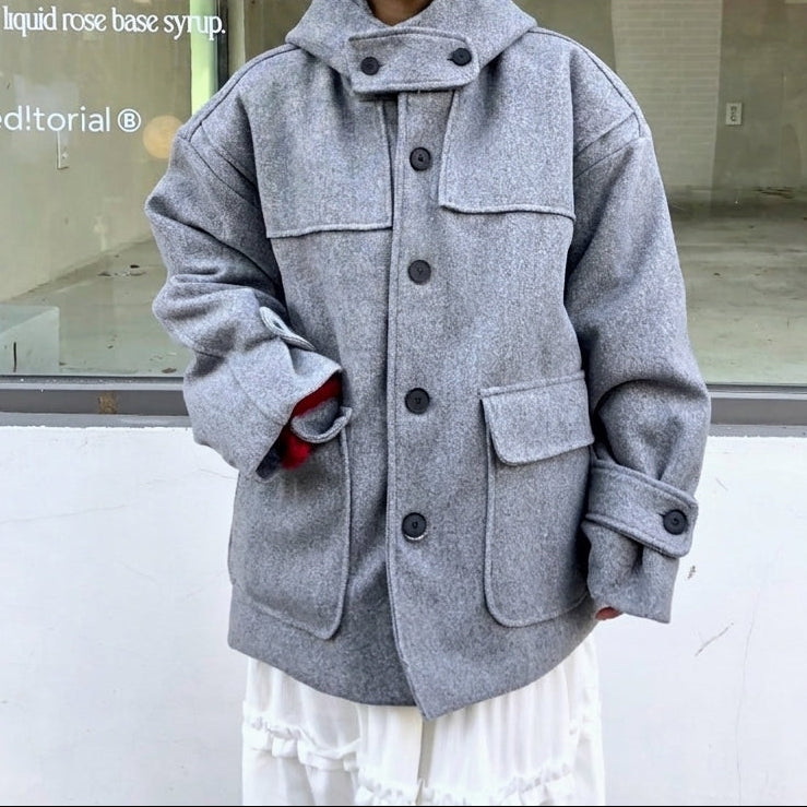 A young woman wearing a light gray oversized wool-blend coat with a high collar and large front pockets, standing in front of a storefront window.