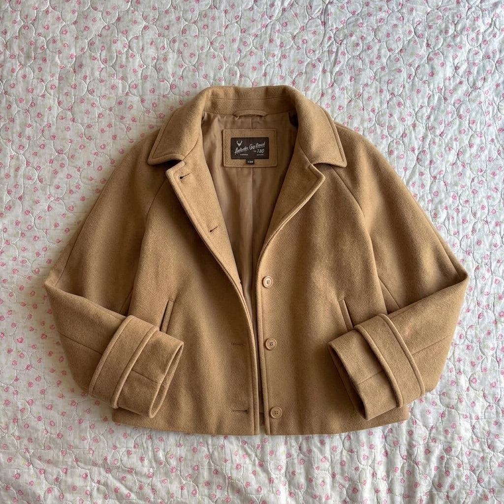 A camel-colored cropped jacket with a collar and button front, laid flat on a pink and white patterned quilted blanket. The brand label inside the collar reads 'Maison Margiela'.