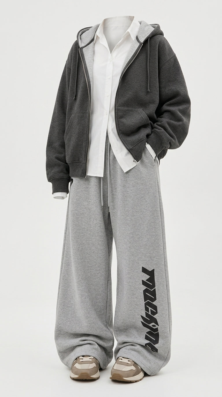 A person wearing a dark gray zip-up hoodie over a white shirt, paired with heather gray sweatpants featuring black vertical text on the lower leg, and beige sneakers, photographed against a light gray studio background.