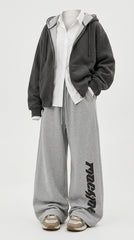 A person wearing a dark gray zip-up hoodie over a white shirt, paired with heather gray sweatpants featuring black vertical text on the lower leg, and beige sneakers, photographed against a light gray studio background.