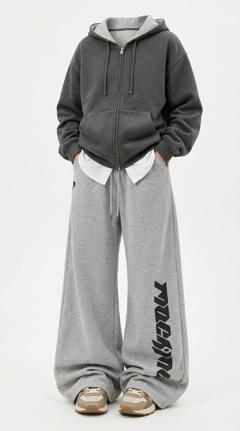 A person wearing an oversized dark gray zip-up hoodie with a white inner collar, paired with wide-leg heather gray sweatpants featuring a black vertical graphic print on the lower leg, and beige sneakers, against a plain white studio background.