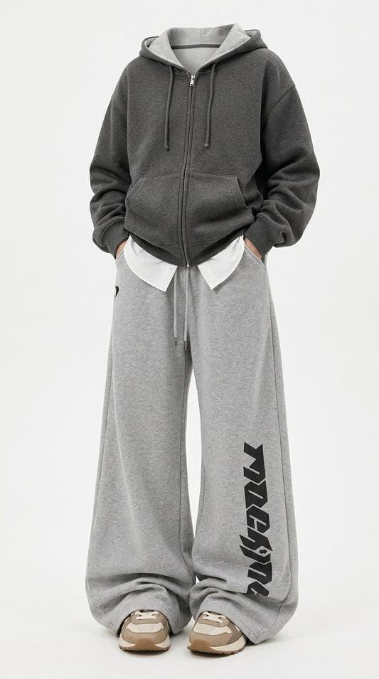 A person wearing an oversized dark gray zip-up hoodie with a white inner collar, paired with wide-leg heather gray sweatpants featuring a black vertical graphic print on the lower leg, and beige sneakers, against a plain white studio background.