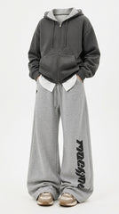 A person wearing an oversized dark gray zip-up hoodie with a white inner collar, paired with wide-leg heather gray sweatpants featuring a black vertical graphic print on the lower leg, and beige sneakers, against a plain white studio background.