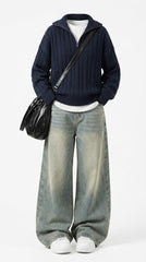 A model wearing a navy blue cable-knit zip-up sweater, wide-leg faded blue jeans, white sneakers, and carrying a black leather shoulder bag, photographed against a plain white studio background.