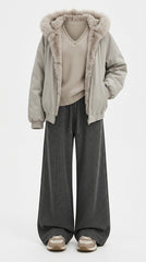 A person wearing a light beige hooded puffer jacket with a fluffy faux-fur trimmed hood, a beige knit sweater, dark gray wide-leg sweatpants, and beige sneakers, shown against a plain light gray studio background.