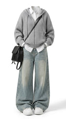 A full-body studio shot of a model wearing a gray zip-up hoodie over a white collared shirt, paired with wide-leg light-wash blue jeans, white sneakers, and a small black crossbody bag, against a plain white background.