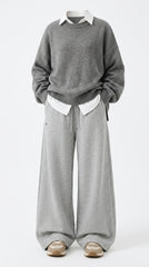 A person wearing a gray knit sweater over a white collared shirt, paired with wide-leg gray sweatpants and beige sneakers, photographed against a plain light gray studio background.