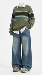 A person wearing an olive green striped sweater with a white collared shirt underneath, wide-leg blue jeans, and gray sneakers, photographed against a plain light background.