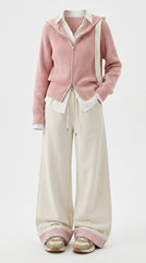 A model wearing a pink ribbed zip-up hoodie over a white collared shirt, paired with wide-leg cream trousers and beige sneakers, shot against a plain white studio background.