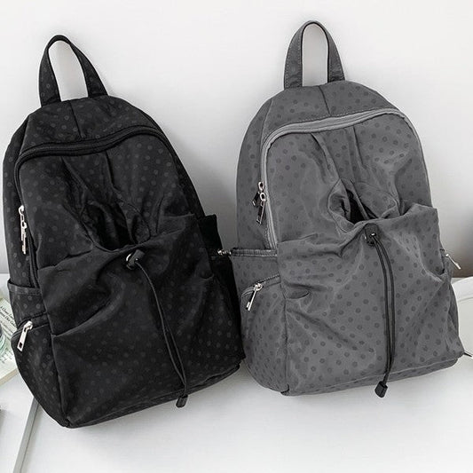 Two backpacks with a subtle polka dot pattern, one black and one gray, displayed side-by-side on a white shelf against a white wall.