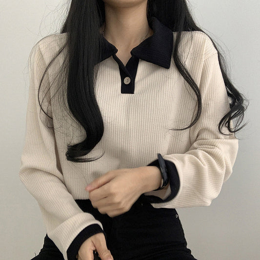 Person wearing a beige ribbed sweater with black collar and cuffs against a plain background