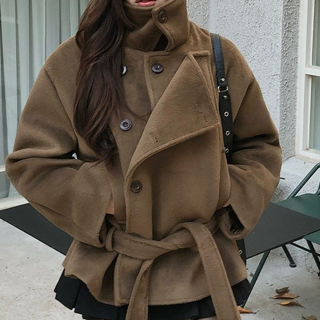 A woman wearing a brown high-neck belted coat with dark tights, standing outdoors in front of a building with windows and a green bench.