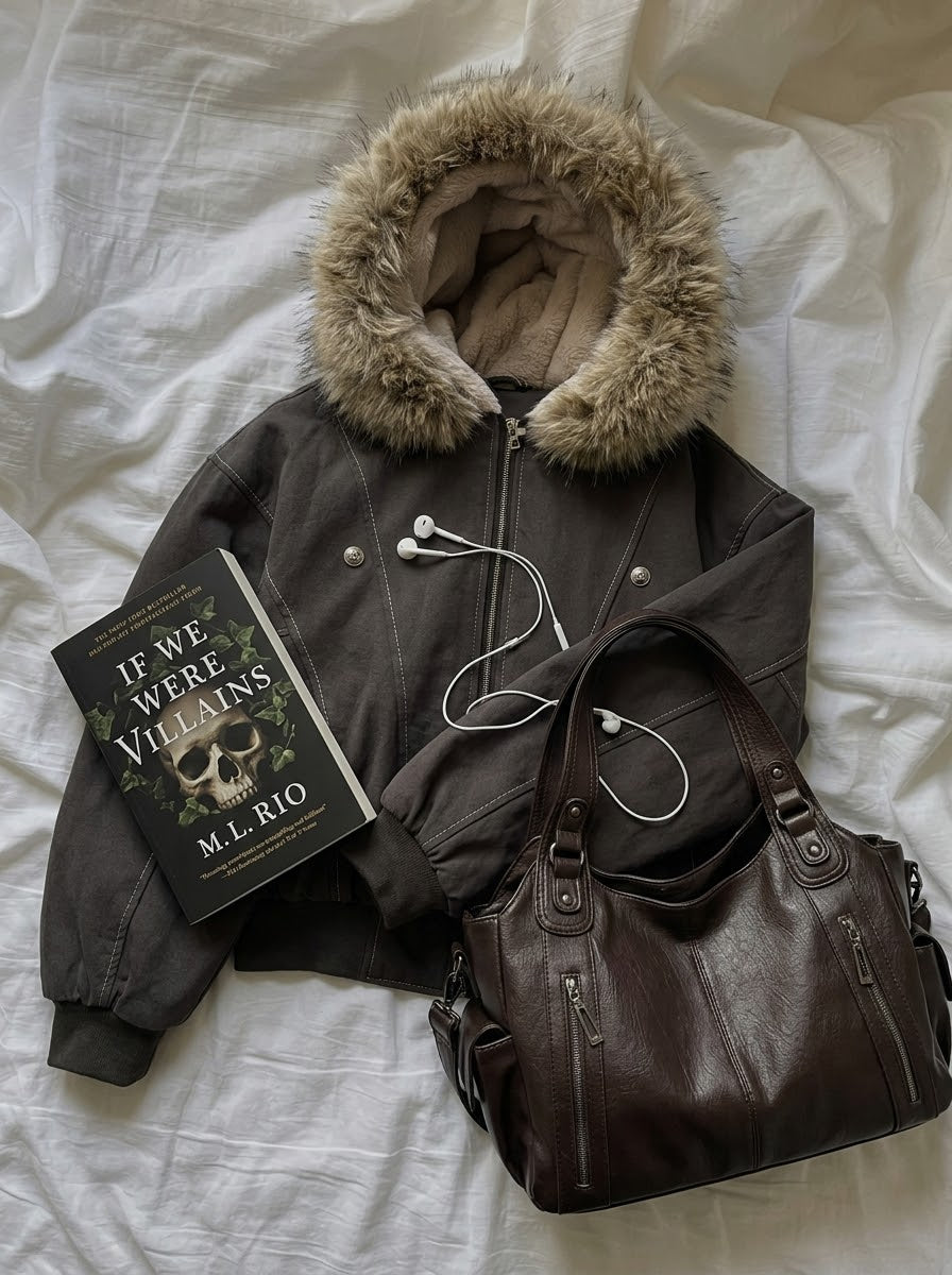 A dark brown faux-fur-trimmed bomber jacket is laid flat on white bedding, styled with a brown leather shoulder bag, a book titled 'If We Were Villains' by M.L.