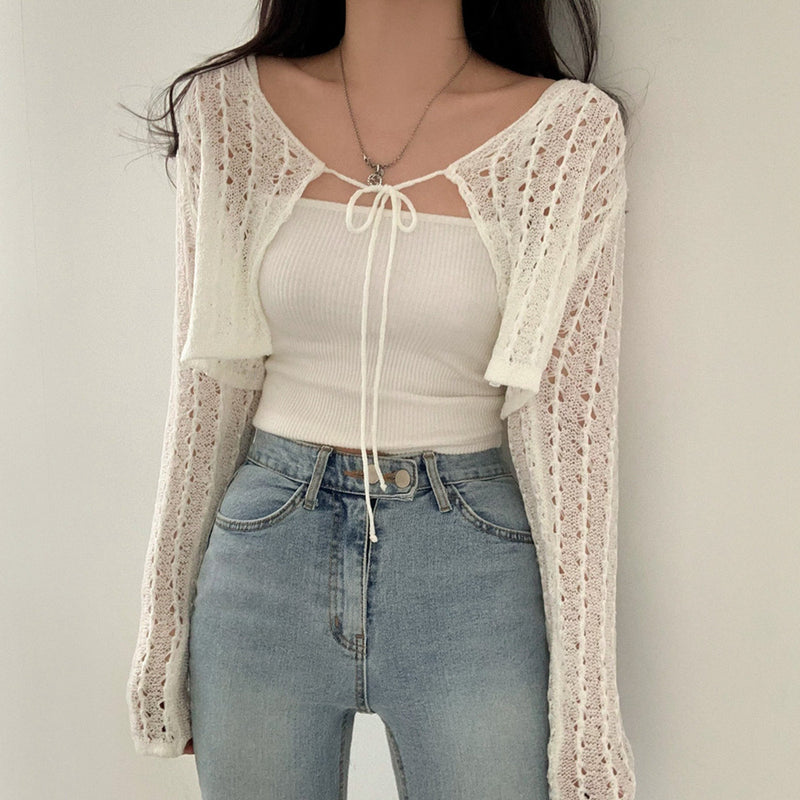 Person wearing a white lace cardigan, beige top, and blue jeans against a plain background