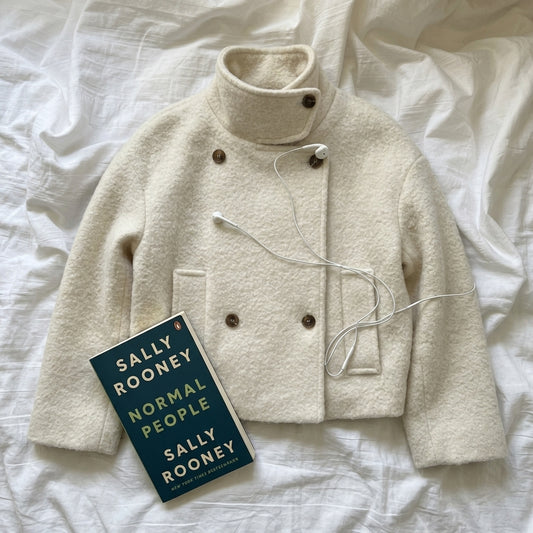 An off-white textured mock-neck jacket with brown buttons, styled on a white wrinkled sheet with a book titled 'Normal People' by Sally Rooney and white wired earphones.