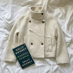 An off-white textured mock-neck jacket with brown buttons, styled on a white wrinkled sheet with a book titled 'Normal People' by Sally Rooney and white wired earphones.
