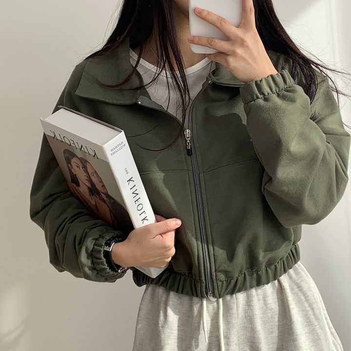 Person wearing a green jacket holding a book and a phone against a plain background
