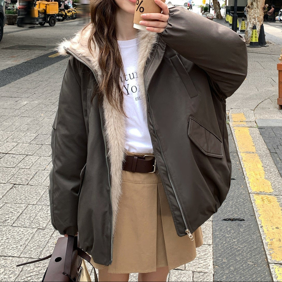 Person wearing a brown jacket with fur lining, white shirt, and beige skirt on a street.