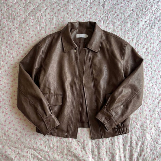 A brown faux-leather jacket with a pointed collar, snap-button front, and flap pockets, laid flat on a light pink and white patterned quilted bedspread.