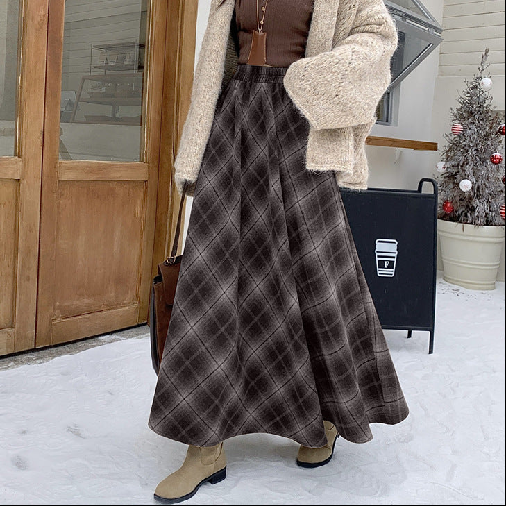Person wearing a plaid skirt and beige coat in front of a wooden door with snow on the ground.